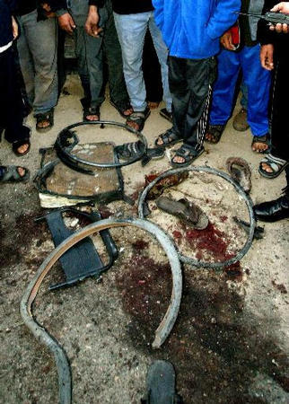 Palestinians gather around pieces of the wheelchair and a blood stain at the scene where an Israeli air strike killed Hamas spiritual leader Ahmed Yassin, Gaza City, March 22, 2004.