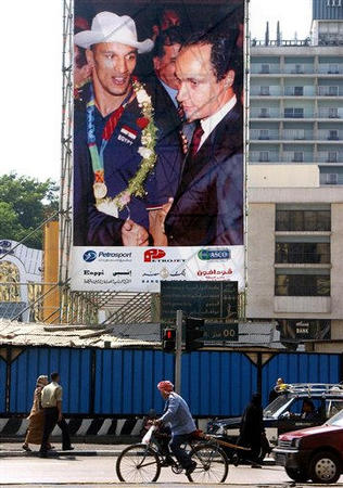 Egyptians pass Friday, a giant billboard showing Gamal Mubarak, Egyptian President Hosni Mubarak's son, right, with wrestling Olympic gold medalist Karam Gaber Ibrahim, pictured when the Egyptian team returned from Athens August 30 with its haul of five medals, including one gold, the country's highest medal count in 56 years, Tahrir Square, Cairo, September 10, 2004.